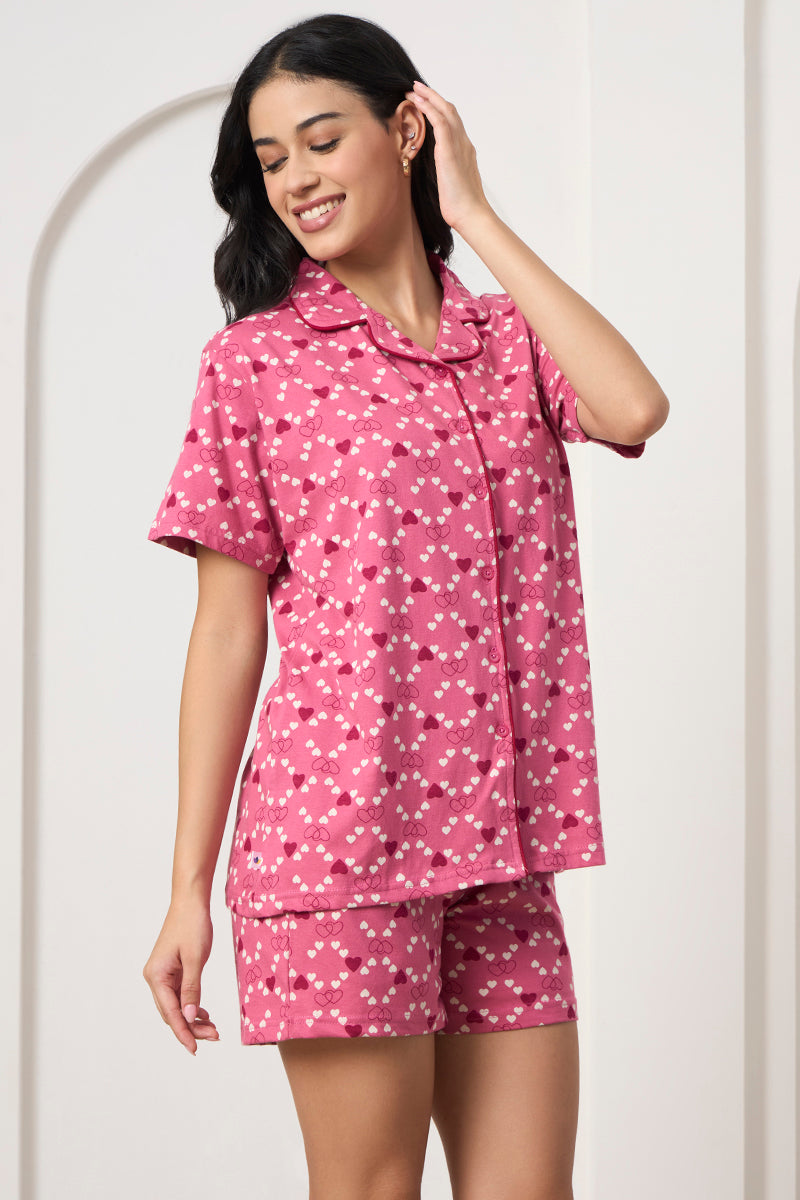 Woman smiling in a pink heart print shirt and shorts set, hand in hair